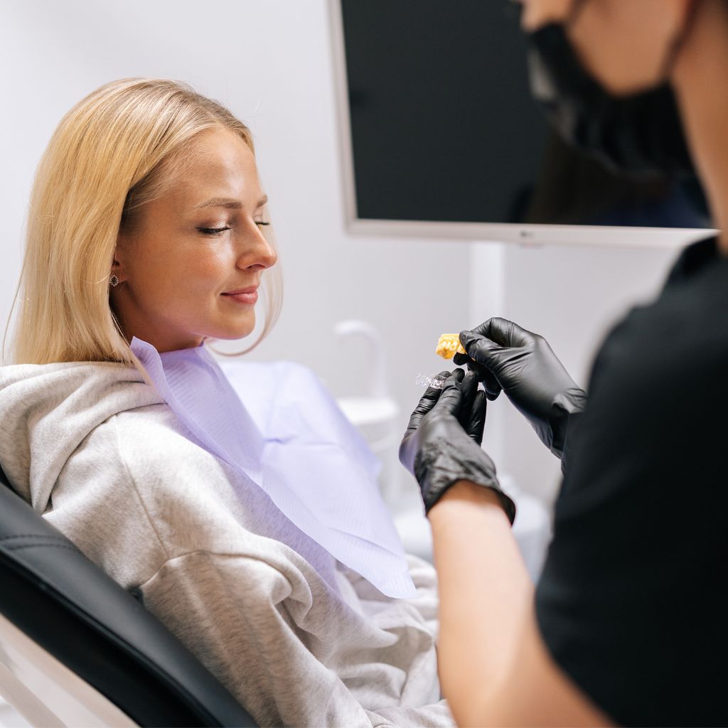 A blonde woman sits comfortably in Dr Theodora Bravis's dentist chair at Thames Dental & Skin Clinic in Thames Ditton, Surrey, near Kingston and Surbiton, as Dr. Bravis demonstrates the Invisalign clear aligners before fitting them, highlighting the clinic's expertise in providing discreet and effective orthodontic treatment.